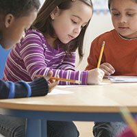 Three children at a classroom table with paper and pencil.